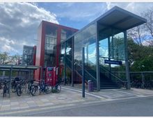 Bahnhof Hoppegarten (Mark) mit moderner Architektur, bestehend aus einem roten Backsteingebäude und einem verglasten Eingangsbereich mit Treppenaufgang. Im Vordergrund befinden sich Fahrradständer mit zahlreichen abgestellten Fahrrädern, umgeben von Bäumen und Himmel mit Wolken im Hintergrund.