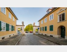 Eine gepflasterte Gasse führt zwischen zwei gelben Gebäuden mit grünen Fensterläden und weißen Sprossenfenstern hindurch. In der Mitte steht ein Torbogen aus Stein mit zwei Skulpturen auf den Pfeilern, dahinter sind Bäume und parkende Autos sichtbar.