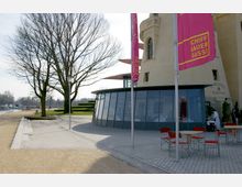 Außenbereich eines modernen Gebäudes mit runden Glasfassaden, gelb verputztem Turm und Werbefahnen mit der Aufschrift „Schiffbauergasse“. Im Vordergrund stehen rote Stühle und Tische eines Restaurants, im Hintergrund befinden sich ein Gehweg, Bäume und ein Wasserlauf.