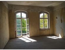 Ein leerer Raum mit abgenutzten, hellgelben Wänden und zwei großen, bogenförmigen Fenstern, von denen eines geöffnet ist und eine kleine Terrasse mit Blick auf Bäume zeigt. Der Boden besteht aus Holzplanken, und Licht fällt durch die Fenster auf den Boden.