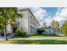 Zweistöckiges Gebäude mit der Aufschrift "Kulturhaus" am Eingang, umgeben von grüner Vegetation und einer gepflasterten Fläche. Rechts schließt ein modernerer Anbau mit großen Glasfenstern an, im Hintergrund Bäume und blauer Himmel.