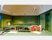 Ein kleiner Sitzungssaal mit grüner Wandvertäfelung, einer gewölbten Decke und modernen Hängelampen. Auf einem grünen Tisch liegen juristische Bücher, Papier und ein Telefon; dahinter stehen Stühle in einer rechteckigen Anordnung.