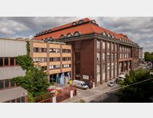 Großes Backsteingebäude mit roten Ziegeldächern und markanten Dachgauben entlang der oberen Etage, daneben ein modernes Bürogebäude mit beigefarbenen Fassadenelementen. Im Vordergrund sind ein Parkplatz, ein rotes Eingangstor sowie Bäume sichtbar.