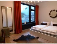 Ein Schlafzimmer mit einem großen Bett, weißen Bettdecken und braunen Kissen. Eine große Fensterfront mit roten Vorhängen dominiert den Raum, ergänzt durch einen Spiegel, einen kleinen Heizkörper, eine Kuhfell-Teppichimitation und hellem Holzboden.