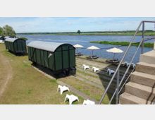 Grüne Eisenbahnwaggons stehen auf einer Wiese am Ufer eines Flusses. Im Vordergrund sind Treppen und weiße Plastikstühle zu sehen, während im Hintergrund Picknicktische mit Sonnenschirmen entlang des Wassers aufgestellt sind.