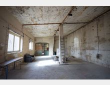 Großer, leerer Innenraum einer alten Industrie- oder Lagerhalle mit rauen, teils abblätternden Wänden und Decke sowie freiliegenden Leitungen und Neonröhren. Links sind mehrere Fenster und eine lange Werkbank, in der Mitte eine Stütze mit angelehnter Leiter, hinten eine grüne Doppeltür und vereinzelte Möbel.