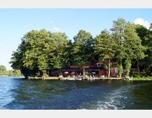 Rotes Backsteingebäude mit Terrasse und bunten Sonnenschirmen am Ufer eines Sees, umgeben von hohen, dicht belaubten Bäumen. Im Vordergrund sind ein Steg und das ruhige Wasser des Sees zu sehen.