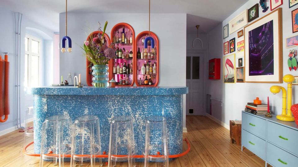 Bright, modern home bar area with a speckled blue counter, clear acrylic bar stools, and hanging pendant lights. Colorful arched wall shelves display bottles behind the bar, with light blue walls, wooden floors, and a gallery wall of framed artwork above a low cabinet.