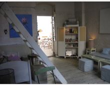 Wohnraum mit Dielenboden und weiß gestrichenen Backsteinwänden. Zu sehen sind ein Hochbett mit Leiter, eine Couch, Regale mit Dekorationsgegenständen und ein Durchgang zu einem helleren Raum mit Holzmöbeln.