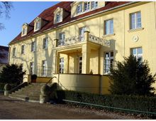 Eine zweistöckige gelbe Villa mit rotem Ziegeldach und mehreren Dachgauben. Das Gebäude hat eine zentrale Treppe, die zu einem überdachten Eingangsbereich mit Säulen und einer Balustrade führt.