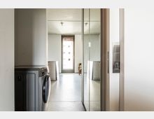 Waschküche mit einer silbernen Waschmaschine links, einem Spiegel an der rechten Wand sowie einem Glas-Schiebetürenelement. Im Hintergrund ist ein Raum mit einem großen Fenster und minimaler Möblierung sichtbar.