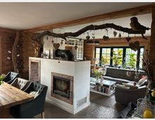 Gemütlicher Wohnraum in einem Holzhaus mit offenen Holzbalken und dekorativen Elementen. Sichtbar sind ein Kamin, eine Sitzecke mit grauem Sofa und ein Esstisch mit Polsterstühlen, umgeben von Pflanzen und Fenstern mit grünen Rahmen.