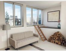 Wohnraum mit minimalistischem Design: Eine beigefarbene Couch steht vor einer weißen Wand, daneben eine Stehlampe. Großflächige Fenster geben den Blick auf eine Terrasse frei. Eine Holzleiter führt zu einer erhöhten Ebene mit gelagertem Brennholz und einer Vase auf einer Ablage.