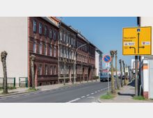 Straße mit historischen Backsteingebäuden auf der linken Seite, teils beschnittenen Bäumen entlang des Bürgersteigs und einem Verkehrsschild mit Wegweisungen zu Zielen wie Neuruppin und Rhinow auf der rechten Seite. Im Hintergrund geparkte und fahrende Fahrzeuge sowie weitere Gebäude.