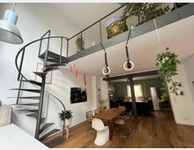 Offener Wohnraum mit moderner Wendeltreppe aus Metall, großem Holztisch und hängenden Turnringen. Im Hintergrund eine Küche mit grüner Wandfarbe und Edelstahlkühlschrank, sowie hängenden Pflanzen und minimalistischem Dekor.