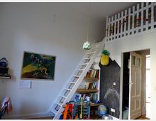 Ein Kinderzimmer mit einer weißen Wand, auf der ein Bild mit Tieren hängt. Eine Leiter führt zu einem Hochbett mit einem weißen Geländer, darunter befindet sich ein Bücherregal und eine Tafelwand mit Zeichnungen. Spielzeug, Bücher und bunte Dekorationen prägen den Raum.