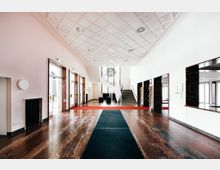 Großzügiger Eingangsbereich mit glänzendem Marmorboden, weißer Kassettendecke und einer zentralen Treppe, die zu einem oberen Stockwerk führt. Auf beiden Seiten befinden sich Türen mit dunklen Marmorumrahmungen, und Tageslicht fällt durch große Fenster ein.