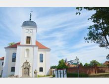 Helle barocke Kirche mit weißer Fassade und rotem Ziegeldach, einem Turm mit Uhr und einer dekorativen Eingangstür. Im Hintergrund sind Bäume, eine rote Backsteinmauer und weitere Gebäude sichtbar.