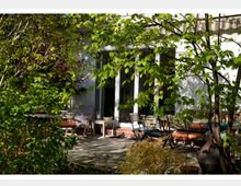 Eine sonnige Terrasse mit Natursteinboden und Gartenmöbeln, umgeben von üppigem Grün und kleinen Bäumen; im Hintergrund eine Hausfassade mit großen Glastüren, die ins Innere führen.