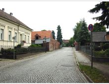 Kopfsteinpflasterstraße in einer ländlichen Wohngegend, gesäumt von gepflegten Einfamilienhäusern mit Vorgärten und Zäunen. Links im Bild steht ein helles Gebäude mit verzierten Fassadenelementen, rechts sind ein Verkehrsschild und einige Nadelbäume zu sehen.