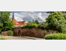 Historische Backsteinmauer mit einer kleinen Durchgangsöffnung, bewachsen mit dichtem Grün und Bäumen; im Hintergrund ein Gebäude mit roten Dachziegeln und einem Glockenturm. Der Vordergrund zeigt eine gepflasterte Straße.