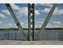 Ein grünes Brückenelement aus Metall mit sichtbaren Nieten steht im Vordergrund, dahinter ein Geländer, ein Fluss und ein bewaldetes Ufer bei klarem Himmel mit wenigen Wolken.