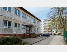 Zweistöckiges Gebäude mit grauer Fassade und Flachdach, auf dessen Vorderseite sich mehrere Fenster und ein überdachter Eingangsbereich mit Treppen und Geländer befinden. Im Hintergrund sind mehrstöckige Wohnhäuser sowie ein Parkplatz mit geparkten Autos zu sehen, daneben stehen Bäume und ein Zaun.