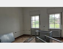 Ein leerer Raum mit zwei großen Fenstern, durch die eine grüne Außenlandschaft sichtbar ist. Die Wände sind weiß gestrichen und teilweise beschädigt, ein Heizkörper befindet sich unter jedem Fenster, und auf dem Boden liegen verlegte Materialien sowie ein Metallrahmen.