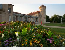 Schloss mit beigem Putz und roten Dächern im spätklassizistischen Stil, flankiert von einem runden Turm und Säulengang. Im Vordergrund ein farbenfrohes Blumenbeet mit Sonnenblumen und anderen Pflanzen, umgeben von gepflegtem Rasen und Gehwegen.