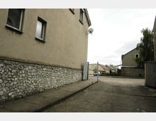 Ein schmaler Gehweg neben einem Gebäude mit verputzter Fassade und einer unteren Steinmauer. Im Hintergrund sind eine ruhige Straße und Häuser mit roten und grauen Dächern sichtbar, umgeben von vereinzelten Bäumen.