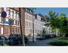 Eine städtische Straßenszene mit mehrstöckigen, dekorativen Wohnhäusern im Altbaustil und parkenden Autos entlang der Straße. Im Vordergrund sind Straßenschilder und Bäume, die den Gehweg säumen, zu sehen.