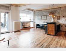 Offener Raum mit rustikalem Holzfußboden, einer Theke aus Holz und schwarzen Akzenten, einer dunkelgrünen Ledercouch und einer Wand mit abgenutzter Putzoptik. Große Fenster lassen viel Tageslicht herein, und von der Decke hängen mehrere Glühlampen.