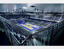 Große, moderne Sporthalle mit einer Basketballspielfläche in der Mitte, umgeben von tribünenartigen Sitzreihen. Die Halle verfügt über ein geräumiges Dach, eine metallene Geländerkonstruktion und zahlreiche Scheinwerfer an der Decke.