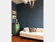 Zimmer mit dunkelblauer Wand, weißer Decke mit sichtbaren Balken und einem Kronleuchter mit farbigen Kerzen. Im Vordergrund steht ein weißes Sofa mit mehreren gemusterten Kissen, daneben ein grüner Metallschrank mit Dekorationen und gerahmten Bildern an der Wand.