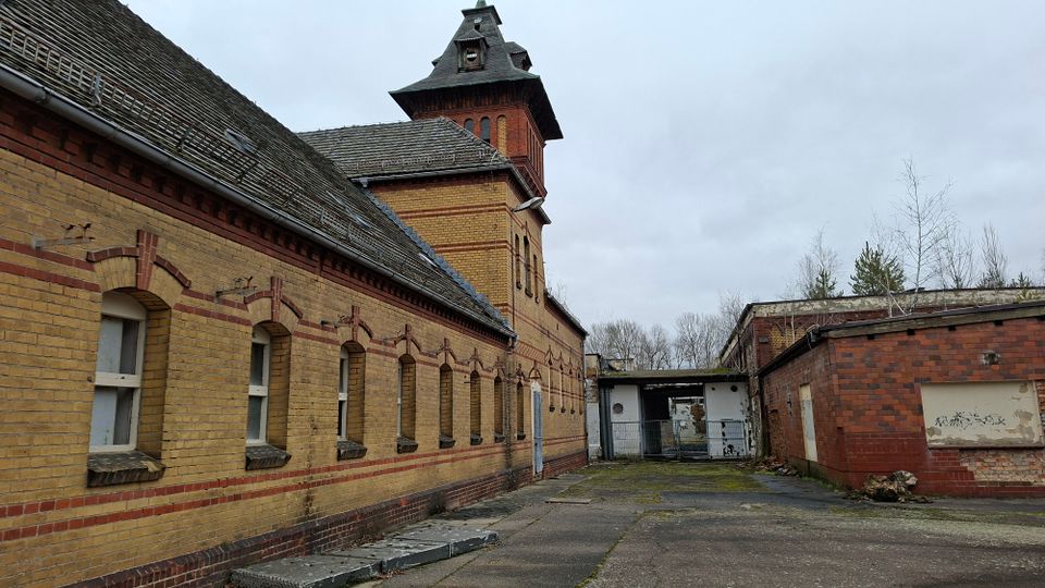Innenhof einer historischen Backsteinanlage mit langem gelb-rotem Ziegelgebäude, Rundbogenfenstern und einem Turm mit steilem Dach. Rechts steht ein niedriger Backsteinbau; dazwischen führt ein überdachter Durchgang zu einem Tor, umgeben von grauem, rissigem Asphalt und vereinzelten Bäumen im Hintergrund.