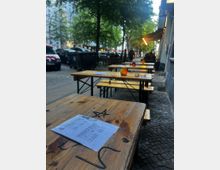 Außenbereich eines Straßencafés mit langen Holztischen und Bänken auf einem Kopfsteinpflaster-Gehweg, auf den Tischen liegen Speisekarten und kleine Kerzen. Entlang der baumbestandenen Straße stehen parkende Autos, und am Gebäude rechts hängt eine ausgefahrene Markise.
