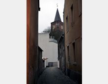 Schmale gepflasterte Gasse mit hohen, einfachen Gebäuden auf beiden Seiten, die zu einem Kirchturm aus rotem Backstein mit Spitzdach und goldenen Zifferblättern führt. Im Hintergrund sind kahle Bäume sichtbar, und ein kleines beleuchtetes Schild ragt links aus einem Gebäude hervor.