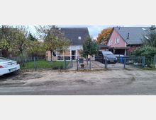 Einfamilienhaus mit Satteldach und heller Fassade, umgeben von einem Garten mit Rasen und Bäumen. Im Vordergrund ein Metallzaun mit Tor, rechts eine Einfahrt mit geparktem Auto.