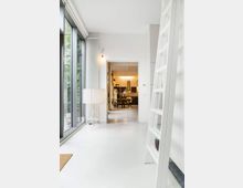 Offener, moderner Innenraum mit weißen Wänden und Boden; links große Fensterscheiben, eine Stehlampe und ein Heizkörper, rechts eine weiße Leiter. Im Hintergrund ein Essbereich mit Tisch, Stühlen und einer schwarz-weißen Küche.