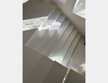 White indoor staircase with wide, rectangular steps leading down to a light gray speckled floor. Sunlight from a nearby opening casts sharp geometric shadows across the steps and adjacent wall.