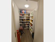 Ein kleiner Abstellraum mit Regalen an beiden Seiten, die mit Ordnern, Büchern, Haushaltsgeräten und verschiedenen Gegenständen gefüllt sind. Der Raum hat Holzfußboden, weiße Wände und eine Deckenlampe in der Mitte.