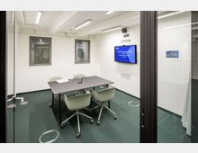 Kleiner Besprechungsraum mit Glaswänden, einem rechteckigen Tisch und vier grünen Bürostühlen auf einem grünen Teppichboden. Eine Wand zeigt einen Bildschirm mit der Aufschrift „Besprechungsraum klein", daneben befinden sich ein Fenster und Lüftungsschlitze.