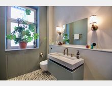 Modernes Badezimmer mit einem rechteckigen Waschbecken aus Marmor, goldenen Wasserhähnen und einem großen Spiegel. Ein Fenster mit einer Pflanze auf der Fensterbank sowie Wandleuchten und gemusterte Bodenfliesen prägen den Raum.