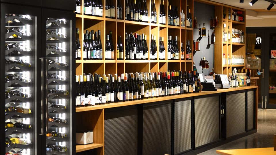 Innenansicht eines Weinladens mit langen Holzregalen voller Weinflaschen entlang der Wand und einer Theke davor. Links stehen beleuchtete Weinkühlschränke mit Glasfront, darüber hängen schwarze Schienenstrahler an der Decke.