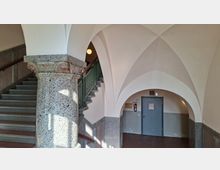 Innenraum eines historischen Treppenhauses mit weißen Kreuzgewölben und rundbogigen Durchgängen, getragen von einer breiten Granitsäule mit verziertem Kapitell. Links führt eine graue Steintreppe mit grünem Geländer nach oben; im Hintergrund befindet sich eine graue Tür in einer gewölbten Nische.