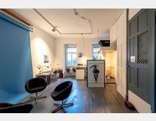 Offener Innenraum eines Ateliers oder Studios mit Holzboden, einer blauen Fotohintergrundrolle an der Wand, mehreren schwarzen Sesseln, Schreibtischen und Hockern. Im Hintergrund befinden sich zwei Fenster mit vergitterten Scheiben, eine kleine Küche und eine Tür, die zu einer Toilette führt.