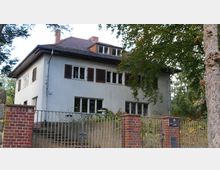 Zweistöckiges Wohnhaus mit weißer Fassade, braunen Fensterläden und einem Satteldach, umgeben von Bäumen. Vor dem Haus befindet sich ein Metallzaun mit gemauerten Backsteinpfeilern.