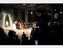 Eine Modenschau in einem modernen Veranstaltungsraum mit Betonwänden und einem gebogenen Durchgang. Zuschauer sitzen entlang des Laufstegs, während Models in heller Kleidung laufen, und Fotografen im Vordergrund die Szene aufnehmen.