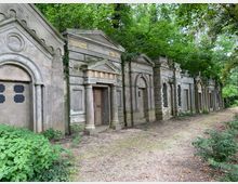 Reihe von historischen Grabmälern auf einem Friedhof, gestaltet mit klassizistischen Elementen wie Säulen, Bögen und Giebeln, umgeben von dichtem Grün und einem unbefestigten Weg im Vordergrund. Die Mauern der Grabstätten zeigen Verwitterungsspuren.