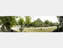 Grüner Dorfplatz mit einem gepflasterten Weg, umgeben von Bäumen und einem grünen Gebäude mit Satteldach und Fenstern. Im Vordergrund sind ein Metallzaun und steinerne Pfosten sichtbar, die den Raum begrenzen.